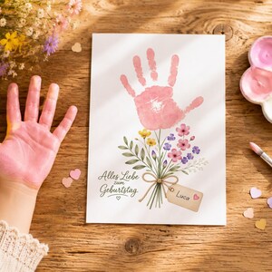 May include: A white rectangular card featuring a pink handprint at the top, which serves as the flower head for a bouquet of illustrated wildflowers. The text "Alles Liebe zum Geburtstag" is printed on the left, and a tag labelled "Luca" is attached.
