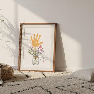 Op de afbeelding: Een ingelijste print met een oranje handafdruk boven een aquarel illustratie van bloemen in een weckpot. De print bevat de tekst "All my love, your Grandbaby" en een klein roze hart. Het frame is van hout.