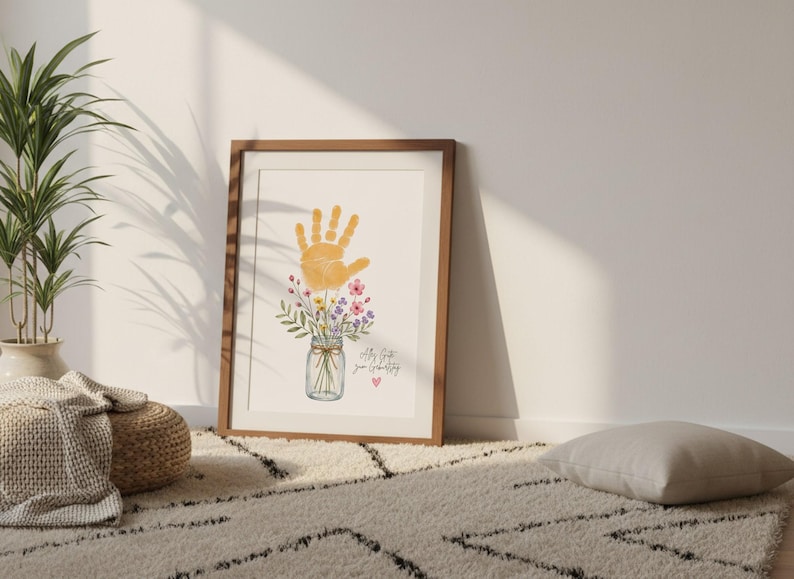 Op de afbeelding: Een ingelijste print met een oranje handafdruk boven een aquarel illustratie van bloemen in een weckpot. De print bevat de tekst "All my love, your Grandbaby" en een klein roze hart. Het frame is van hout.