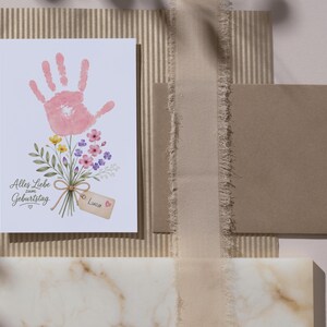 May include: A rectangular greeting card featuring a pink handprint illustration transformed into a flower blossom atop a bouquet of colourful wildflowers. The text reads Alles Liebe zum Geburtstag with a small tag labelled Luca and a heart icon.