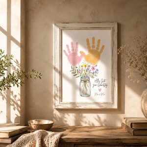 Op de afbeelding: Ingelijste kunstdruk met roze en gele handafdrukken boven een aquarelpot met bloemen. De print bevat de tekst "Alles Gute zum Geburtstag" en "Mama & Papa" in zwart schrift, tegen een neutrale achtergrond.