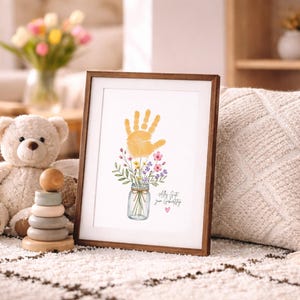 Op de afbeelding: Een ingelijste print met een gele handafdruk boven een aquarel illustratie van bloemen in een pot. De print heeft een bruine houten lijst en de tekst "Let's Get Your Creativity". Een teddybeer en een houten stapelspeelgoed staan op de achtergrond.