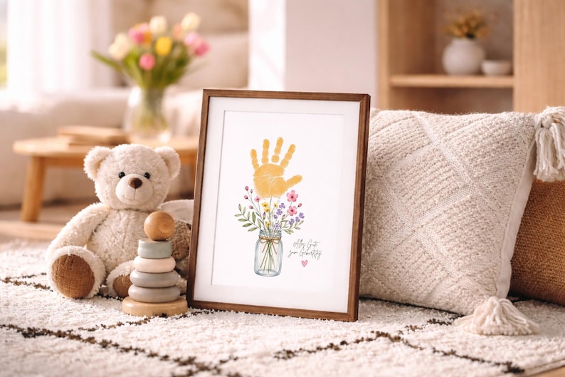 Op de afbeelding: Een ingelijste print met een gele handafdruk boven een aquarel illustratie van bloemen in een pot. De print heeft een bruine houten lijst en de tekst "Let's Get Your Creativity". Een teddybeer en een houten stapelspeelgoed staan op de achtergrond.