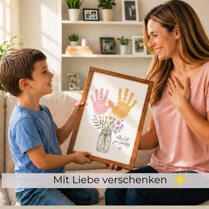 Op de afbeelding: Een ingelijst kunstwerk met roze en gele handafdrukken en een bloemenboeket in een glazen pot. Het kunstwerk wordt door een kind aan een volwassene gepresenteerd. De tekst "Mit Liebe verschenken" staat onderaan.