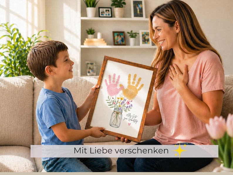 Op de afbeelding: Een ingelijst kunstwerk met roze en gele handafdrukken en een bloemenboeket in een glazen pot. Het kunstwerk wordt door een kind aan een volwassene gepresenteerd. De tekst "Mit Liebe verschenken" staat onderaan.