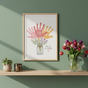 Op de afbeelding: Ingelijste kunstdruk met een boeket van roze en gele handafdrukken, gerangschikt in een weckpot met bloemen. De tekst "all that you create" staat eronder. De print wordt op een houten plank gepresenteerd.