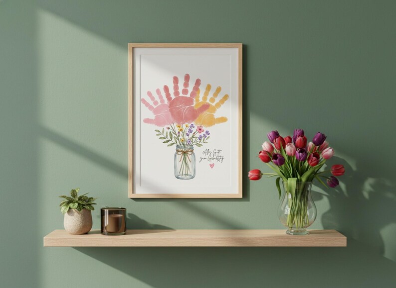 Op de afbeelding: Ingelijste kunstdruk met een boeket van roze en gele handafdrukken, gerangschikt in een weckpot met bloemen. De tekst "all that you create" staat eronder. De print wordt op een houten plank gepresenteerd.