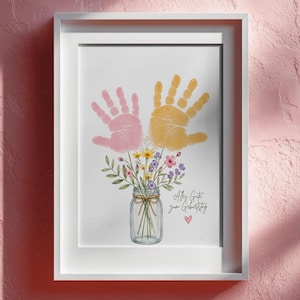 Op de afbeelding: Ingelijste print met roze en gele handafdrukken boven een aquarel illustratie van een bloemenboeket in een weckpot. De print bevat de tekst "Alles Gute zum Geburtstag" en een klein roze hart.