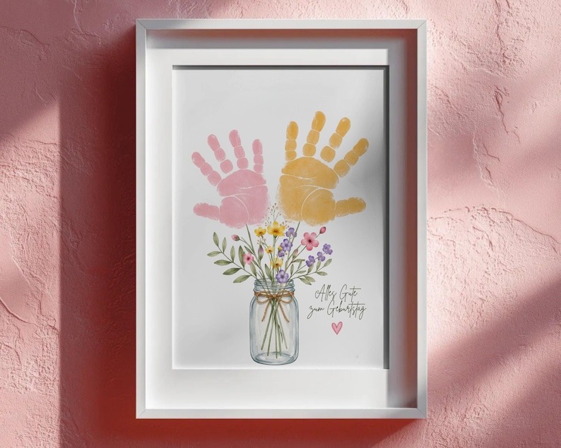 Op de afbeelding: Ingelijste print met roze en gele handafdrukken boven een aquarel illustratie van een bloemenboeket in een weckpot. De print bevat de tekst "Alles Gute zum Geburtstag" en een klein roze hart.