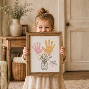 Op de afbeelding: Een houten lijst met twee handafdrukken, roze en geel, boven een bloemstuk in een pot. De tekst luidt "All my love, your Grandbaby". De lijst is van hout.