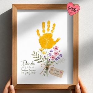 May include: A framed print featuring a yellow handprint positioned above a watercolour bouquet of small pink, yellow, and purple flowers. The text reads: Danke für all die Tage voller Lachen, Lernen und Geborgenheit, with a tag labelled Deine Emma.