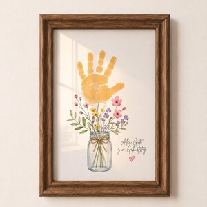 Op de afbeelding: Een ingelijste print met een gele handafdruk boven een pot gevuld met kleurrijke bloemen. De pot is vastgebonden met touw. De print bevat de tekst "Alles Gute zum Geburtstag" en een roze hart.