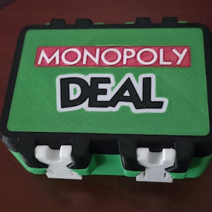 May include: A green Monopoly Deal card game box with black trim and white clasps. The box features the words "MONOPOLY DEAL" in white and red lettering.
