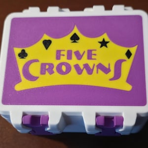 May include: A white and purple Five Crowns card game box. The lid features a yellow crown design with card suit symbols and the game's name in purple. The box has purple clasps.