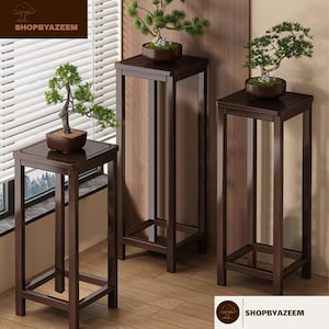 以下が含まれることがあります： 高さの異なる3つの濃い茶色の木製プラントスタンド。それぞれ正方形の天板と下の棚が付いています。各スタンドには、茶色の鉢植えの盆栽が入っています。スタンドは木の床にあります。