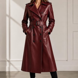 May include: A long, double-breasted trench coat in a deep burgundy color, crafted from a smooth material. The coat features a wide notched collar, shoulder epaulets, long sleeves with buckled cuffs, and a matching waist belt with a rectangular buckle.