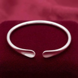 May include: A polished silver-tone cuff bracelet with a smooth, rounded surface and an open design. The bracelet features a simple, continuous band that curves into two rounded, slightly flattened ends, creating a clean and reflective metallic finish.