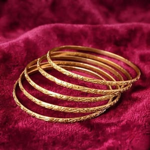 Puede incluir: Un conjunto de cinco brazaletes finos de color dorado, cada uno con un patrón repetitivo, delicado y texturizado en la superficie exterior. Las pulseras circulares están apiladas ligeramente superpuestas, mostrando un acabado metálico.