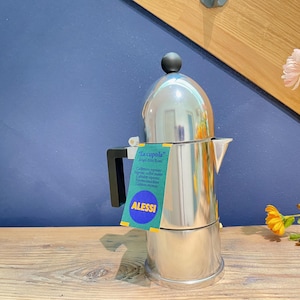 Può includere: Una caffettiera espresso in acciaio inossidabile con manico nero e pomello nero sulla parte superiore. Il prodotto ha un'etichetta con la scritta "La cupola" e "ALESSI". La caffettiera è su una superficie di legno con sfondo blu.