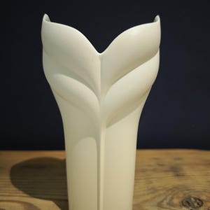 May include: A white ceramic vase with a distinctive leaf design. The vase has a tall, slender body that widens at the top, with two curved, petal-like shapes. The vase is set against a dark blue background and a wooden surface.