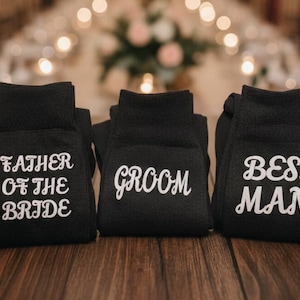 Pode incluir: Três pares de meias pretas com texto branco. Um par diz "FATHER OF THE BRIDE", outro "GROOM" e o terceiro "BEST MAN". As meias estão dobradas e dispostas sobre uma superfície de madeira.