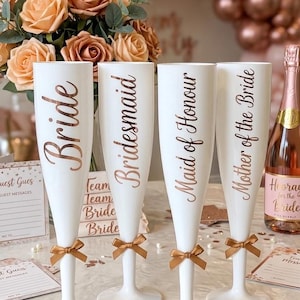 Puede incluir: Cuatro copas de champán blancas con letras en oro rosa que dicen Bride, Bridesmaid, Maid of Honour y Mother of the Bride. Cada copa tiene un pequeño lazo dorado metálico atado alrededor del tallo, colocadas sobre una superficie plana.