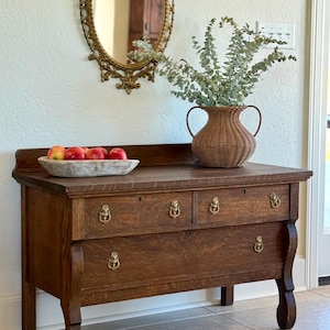 May include: A vintage wooden dresser with three drawers and brass hardware. A woven vase with greenery sits on top, along with a wooden bowl of red apples. An ornate oval mirror hangs on the wall above.