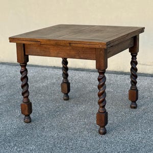 May include: A square, antique wooden table with a dark brown finish. The table features a rectangular top and four legs with a twisted, spiral design. The table is approximately 24 inches wide and 24 inches long.