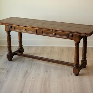 May include: A long, rectangular wooden console table with a dark brown finish. The table features a flat top, two small drawers with round knobs, and four turned legs connected by a horizontal stretcher bar near the base.