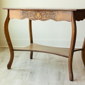 May include: A brown wooden console table featuring curved legs and a lower shelf. The front apron displays intricate carved leaf patterns surrounding a central oval shape. The table has a rectangular top and a smooth, polished wood finish.