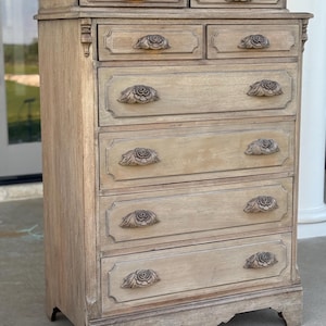 May include: A tall, light brown wooden dresser with seven drawers and ornate rose-shaped drawer pulls. The dresser has a rectangular shape with a small top section and decorative molding.