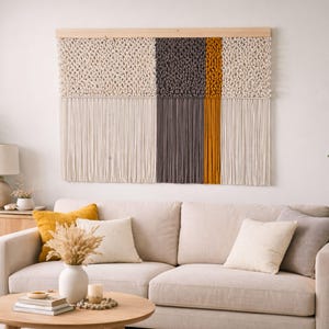 Puede incluir: Un gran tapiz de macramé con una barra de madera. El diseño presenta secciones en crema, gris y amarillo mostaza. Debajo de las secciones tejidas hay flecos color crema. El tapiz se exhibe sobre un sofá beige.