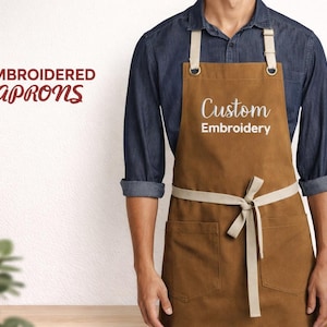 Puede incluir: Un delantal marrón con una correa para el cuello y una corbata en la cintura de color crema. El delantal tiene dos bolsillos delanteros y las palabras "Custom Embroidery" están escritas en escritura blanca. Las palabras "Embroidered Aprons" están en la esquina superior izquierda.