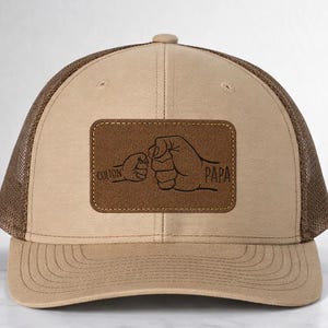 Pode incluir: Boné de caminhoneiro bege e castanho com costas em malha. A frente apresenta um patch de couro castanho com as palavras "Colton" e "Papa" e um design de punho.