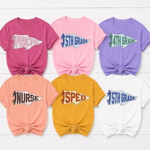 Könnte beinhalten: Sechs kurzärmelige T-Shirts in verschiedenen Farben wie Magenta, Hellrosa, Lila, Pfirsich, Senfgelb und Weiß. Jedes T-Shirt zeigt eine Wimpel-Grafik mit dem Text PRINCIPAL, 5TH GRADE, 4TH GRADE, NURSE oder SPED auf der Vorderseite.