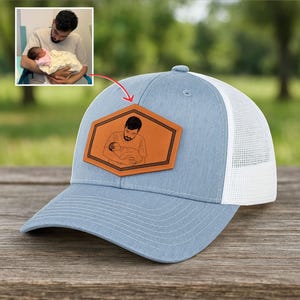 Könnte beinhalten: Hellblaue Trucker-Cap mit weißem Mesh-Rücken und einem braunen Lederaufnäher. Der Aufnäher zeigt eine Strichzeichnung einer Person, die ein Baby hält. Die Kappe hat einen gebogenen Schirm und einen Knopf oben.