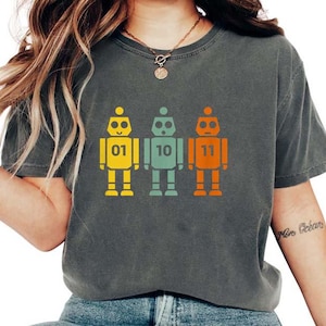 Könnte beinhalten: Dunkelgraues T-Shirt mit drei Retro-Roboter-Grafiken in Gelb, Türkis und Orange. Jeder Roboter hat eine Nummer auf der Brust: 01, 10 und 11. Die Roboter haben ein Vintage-Pixel-Design.