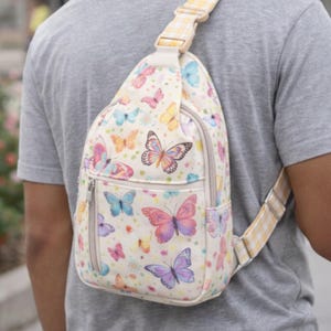 May include: A cream-colored sling bag adorned with colorful butterflies in shades of pink, blue, purple, and orange. The bag features a zippered pocket and a yellow and white checkered strap. The bag is worn over the shoulder.