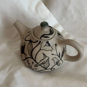Op de afbeelding: Een keramische theepot met een gespikkelde beige afwerking. De theepot heeft een zwart bloemen- en vogelontwerp. Het deksel heeft een kleine groene knop. Handvat en tuit zijn gebogen. Een decoratief item voor de keuken.