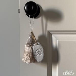以下が含まれることがあります： 明るいベージュのタッセルとシルバー色の楕円形のタグが付いた装飾的なドアタッセル。タグには筆記体フォントで刺繍された文字「R」が施されています。金色の留め金がタッセルとタグを白いコードに接続しています。