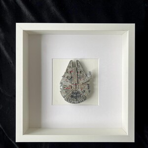 May include: A framed model of the Millennium Falcon starship. The model is grey with red and black details, set against a white background within a white frame. The frame is displayed on a black velvet surface.