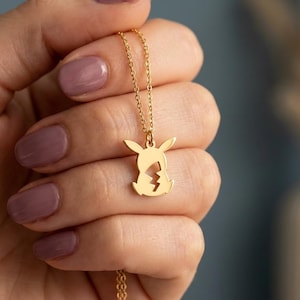 Könnte beinhalten: Eine goldfarbene Anhängerkette mit einem Pikachu-Design. Der Anhänger ist eine Silhouette der Figur mit einem Blitzdetail. Die Kette wird in einer Hand mit mauvefarbenem Nagellack gehalten.