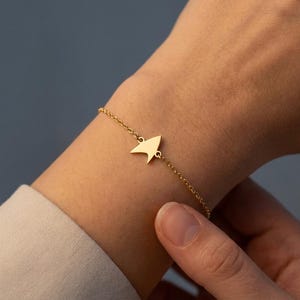 May include: A gold-coloured bracelet featuring a delicate chain and a central charm shaped like a stylised delta insignia. The metal charm has a smooth, polished finish and is attached to the chain links on both sides, creating a simple, refined look.