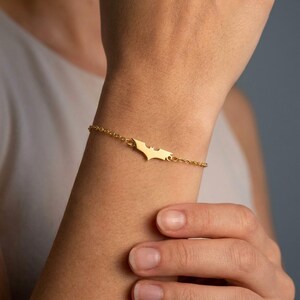 May include: A gold-toned metal bracelet featuring a central cutout charm shaped like a bat silhouette. The charm is attached to a delicate, thin link chain that wraps around the wrist, secured with a small clasp mechanism visible on the side.