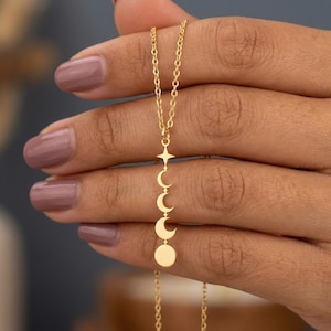 May include: A gold-colored necklace featuring a star and moon phase pendant. The pendant showcases a star, followed by crescent moons in different phases, and a full moon at the bottom. The chain is delicate and the necklace is held by a hand.