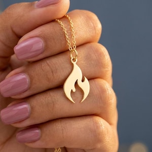 May include: A gold-colored necklace featuring a flame pendant. The pendant has a smooth, polished surface and is suspended from a delicate chain. The necklace is held in a hand with pink nail polish.