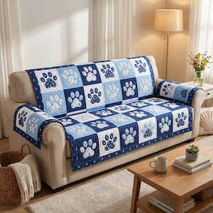 Può includere: Una coperta trapuntata blu e bianca per divano con un motivo a impronte di zampe. La coperta è composta da blocchi quadrati in tonalità di blu e bianco, con impronte di zampe al centro di ogni quadrato. La coperta è drappeggiata su un divano beige.