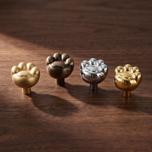 May include: Four cabinet knobs shaped like animal paws, arranged in a row. The knobs feature different metallic finishes, including matte gold, antique bronze, polished chrome, and polished gold, each with a central pad and four toe details.