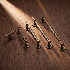 May include: A set of five brass-toned metal cabinet pulls and a knob, each featuring a textured, hammered surface finish. The hardware pieces vary in length, with each handle supported by two circular mounting bases and the knob by a single base.