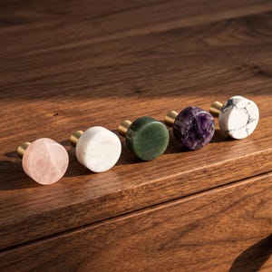 Puede incluir: Cinco pomos circulares para armarios con bases de metal dorado. Los pomos presentan caras de piedra distintas: cuarzo rosa pálido, mármol blanco sólido, jade verde intenso, amatista morada oscura y mármol blanco con vetas grises.